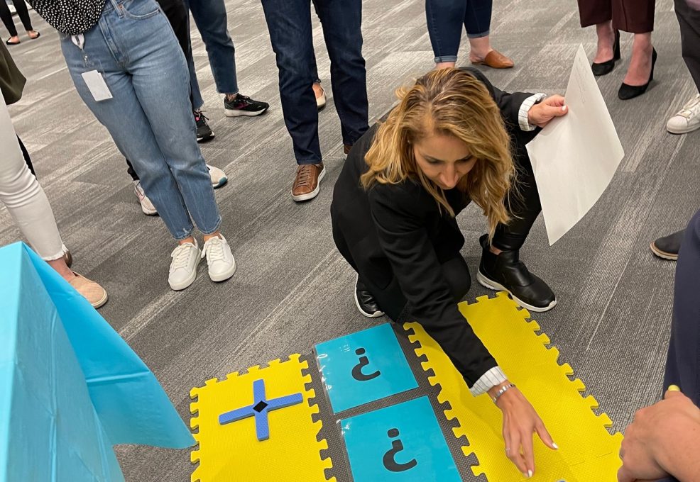 A participant carefully places a marker on a large tic-tac-toe style game board during a Foodbank Showdown team building event. The surrounding group watches intently as the competition unfolds.