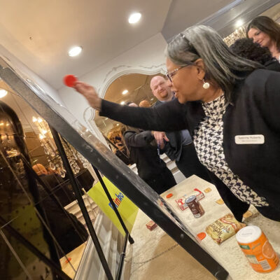 A participant enthusiastically throws a red disc onto a game board during the Foodbank Showdown team building event. In the background, grocery items are visible, showcasing the event's purpose of gathering goods for charity. thumbnail