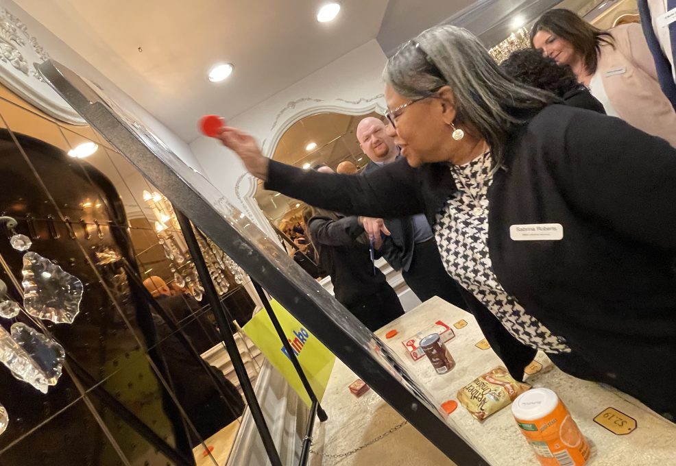 A participant enthusiastically throws a red disc onto a game board during the Foodbank Showdown team building event. In the background, grocery items are visible, showcasing the event's purpose of gathering goods for charity.