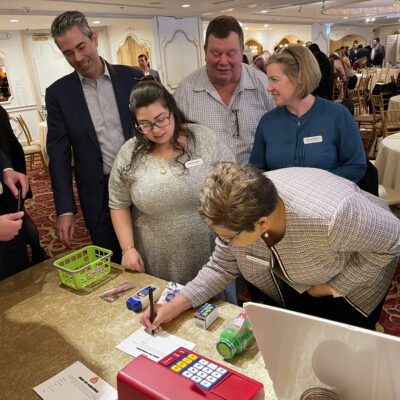 Participants gather around a table engaging in a fun grocery-themed challenge during the Foodbank Showdown team building event. A red toy cash register and mini grocery items are on the table, emphasizing the game's interactive and charitable aspect. thumbnail