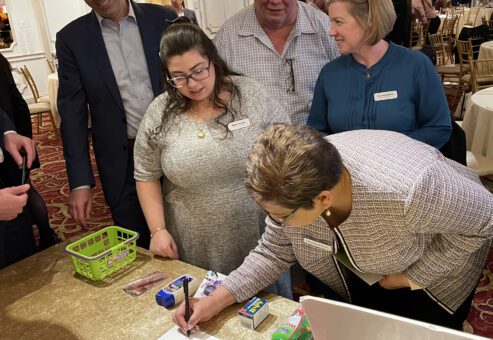 Participants gather around a table engaging in a fun grocery-themed challenge during the Foodbank Showdown team building event. A red toy cash register and mini grocery items are on the table, emphasizing the game's interactive and charitable aspect.