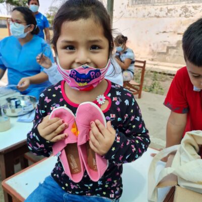 A young girl wearing a colorful mask smiles as she holds a pair of pink TOMS shoes that she received through the Paint Soles for Souls charitable team building event. In the background, healthcare workers assist other children. thumbnail