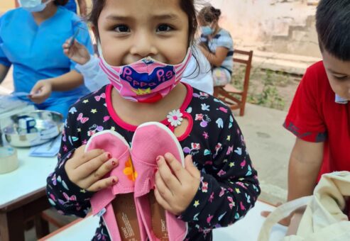 A young girl wearing a colorful mask smiles as she holds a pair of pink TOMS shoes that she received through the Paint Soles for Souls charitable team building event. In the background, healthcare workers assist other children.