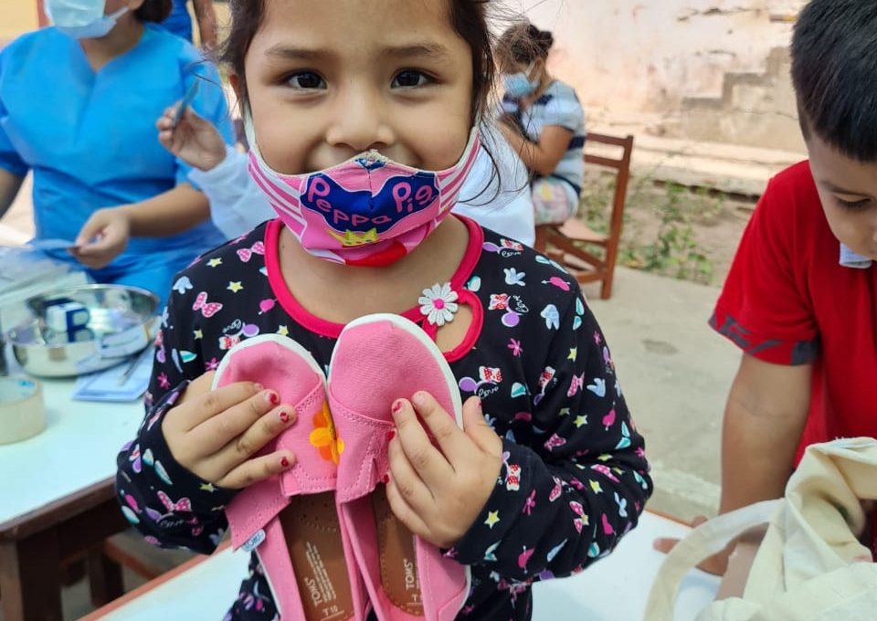 A young girl wearing a colorful mask smiles as she holds a pair of pink TOMS shoes that she received through the Paint Soles for Souls charitable team building event. In the background, healthcare workers assist other children.