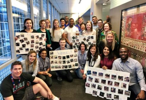 A large group of team members posing together in a hallway, proudly displaying their collage boards filled with Polaroid photos from a scavenger hunt team building event. The boards are labeled with team names and showcase the creative challenges completed during the activity. The group smiles in celebration, in a fun and energetic atmosphere.