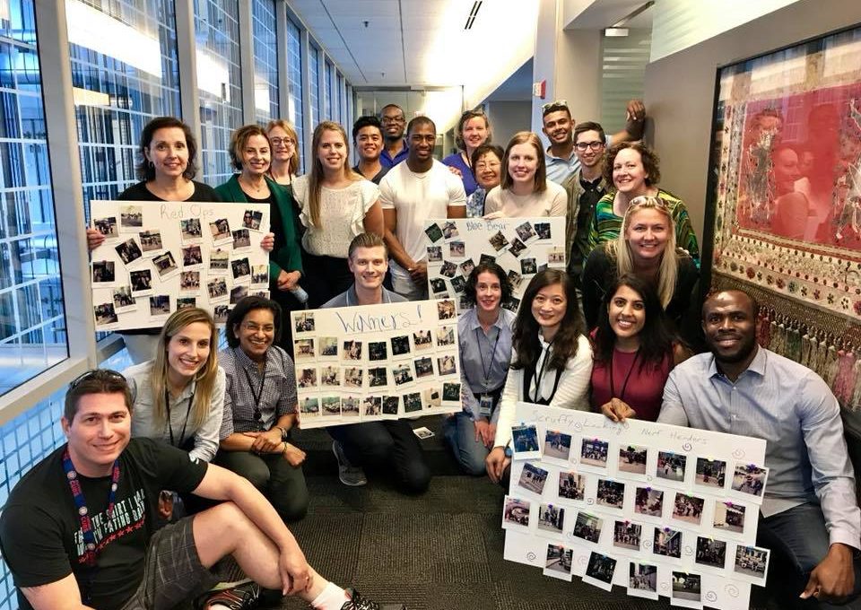 A large group of team members posing together in a hallway, proudly displaying their collage boards filled with Polaroid photos from a scavenger hunt team building event. The boards are labeled with team names and showcase the creative challenges completed during the activity. The group smiles in celebration, in a fun and energetic atmosphere.