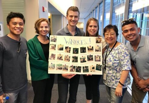 A group of six team members proudly display their winning collage of Polaroid photos from a scavenger hunt. They pose together in a hallway, smiling and celebrating their successful completion of fun and creative challenges, with the board labeled 'Winners!' showcasing the captured moments from the team building activity.