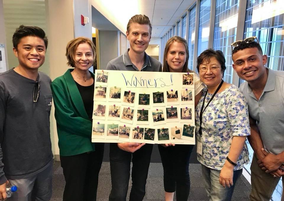A group of six team members proudly display their winning collage of Polaroid photos from a scavenger hunt. They pose together in a hallway, smiling and celebrating their successful completion of fun and creative challenges, with the board labeled 'Winners!' showcasing the captured moments from the team building activity.