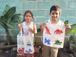 Two smiling children proudly hold up decorated tote bags they received through the Paint Soles for Souls charitable team building event. The bags feature colorful designs of dinosaurs, trains, and sailboats, hand-painted by participants.