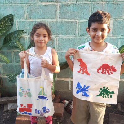 Two smiling children proudly hold up decorated tote bags they received through the Paint Soles for Souls charitable team building event. The bags feature colorful designs of dinosaurs, trains, and sailboats, hand-painted by participants. thumbnail