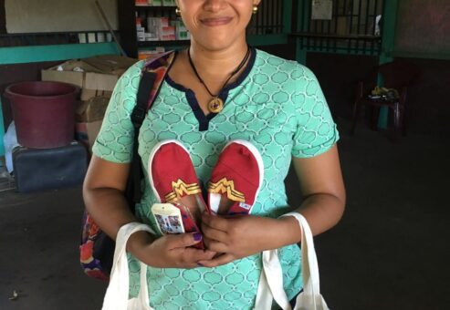 A smiling woman holds a pair of red, hand-painted TOMS shoes featuring a Wonder Woman design, along with decorated tote bags, which were donated through the Paint Soles for Souls charitable team building event.