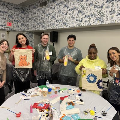 Participants proudly display their painted TOMS shoes and tote bags during the Paint Soles for Souls team building event, showcasing vibrant designs including a cat and a flower. thumbnail