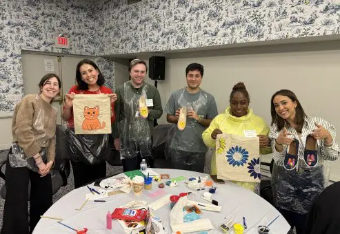 Participants proudly display their painted TOMS shoes and tote bags during the Paint Soles for Souls team building event, showcasing vibrant designs including a cat and a flower.