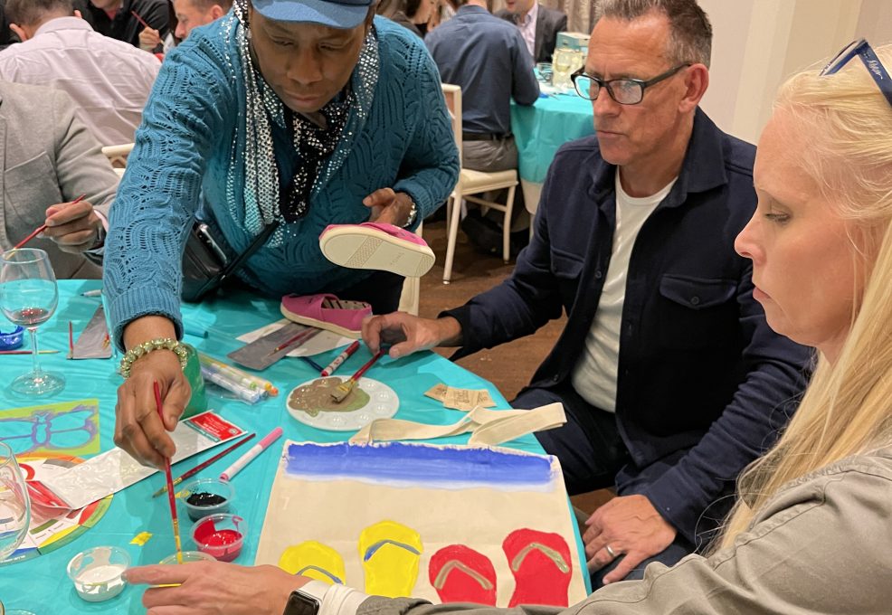 Participants work together to paint TOMS shoes and tote bags during the Paint Soles for Souls charitable team building event. One participant paints a tote bag with colorful flip-flop designs while others decorate shoes with intricate patterns.
