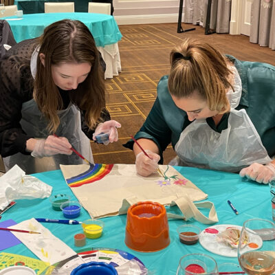 Two women wearing aprons are focused on painting tote bags with colorful designs, including rainbows and flowers, during the Paint Soles for Souls charitable team building event. thumbnail