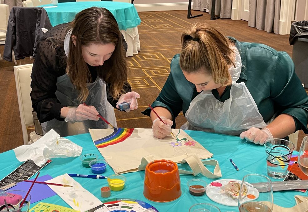 Two women wearing aprons are focused on painting tote bags with colorful designs, including rainbows and flowers, during the Paint Soles for Souls charitable team building event.