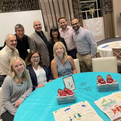 A group of participants smiles as they display their hand-painted TOMS shoes and tote bags during the Paint Soles for Souls charitable team building event. The red shoes and creatively designed bags will be donated to children in South America. thumbnail
