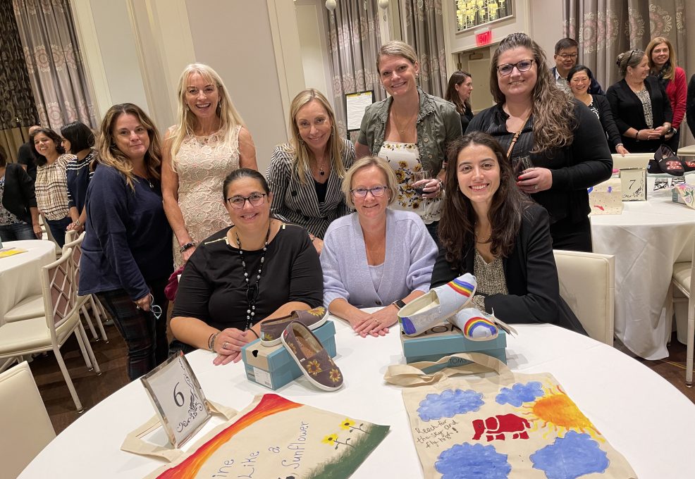 A group of participants smiles as they showcase their hand-painted TOMS shoes and tote bags during the Paint Soles for Souls charitable team building event. The colorful designs, including sunflowers and rainbows, are ready to be donated.