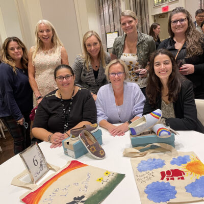A group of participants smiles as they showcase their hand-painted TOMS shoes and tote bags during the Paint Soles for Souls charitable team building event. The colorful designs, including sunflowers and rainbows, are ready to be donated. thumbnail
