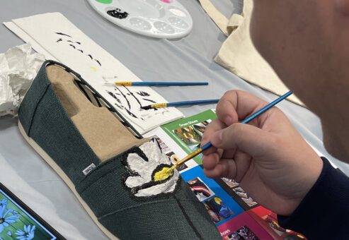 A participant carefully paints a white flower on a pair of green TOMS shoes during the Paint Soles for Souls charitable team building event. The table is covered with art supplies, including paintbrushes, a palette, and design inspiration cards.