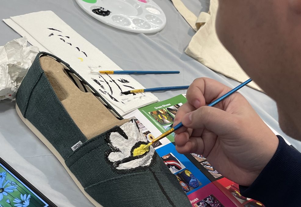 A participant carefully paints a white flower on a pair of green TOMS shoes during the Paint Soles for Souls charitable team building event. The table is covered with art supplies, including paintbrushes, a palette, and design inspiration cards.