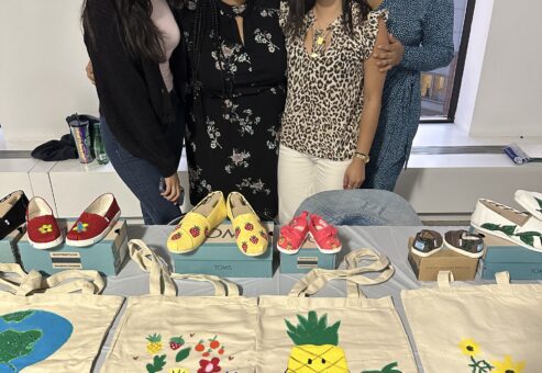 Four smiling participants stand behind a display of hand-painted TOMS shoes and decorated tote bags during the Paint Soles for Souls charitable team building event. The shoes and bags feature colorful designs, including flowers, pineapples, and strawberries, which will be donated to children in need in South America.