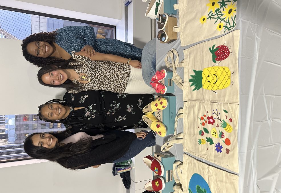 Four smiling participants stand behind a display of hand-painted TOMS shoes and decorated tote bags during the Paint Soles for Souls charitable team building event. The shoes and bags feature colorful designs, including flowers, pineapples, and strawberries, which will be donated to children in need in South America.