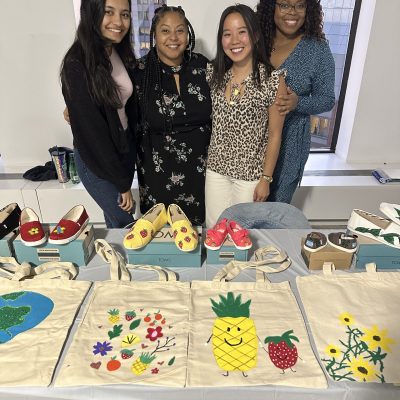 Four smiling participants stand behind a display of hand-painted TOMS shoes and decorated tote bags during the Paint Soles for Souls charitable team building event. The shoes and bags feature colorful designs, including flowers, pineapples, and strawberries, which will be donated to children in need in South America. thumbnail