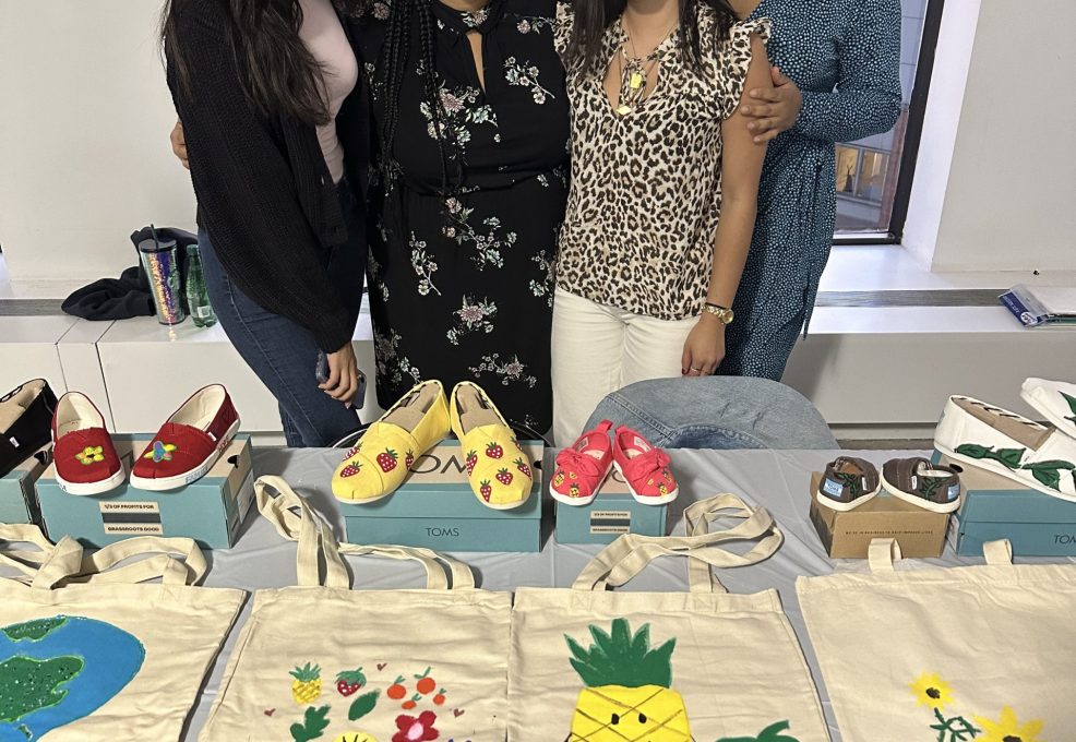 Four smiling participants stand behind a display of hand-painted TOMS shoes and decorated tote bags during the Paint Soles for Souls charitable team building event. The shoes and bags feature colorful designs, including flowers, pineapples, and strawberries, which will be donated to children in need in South America.