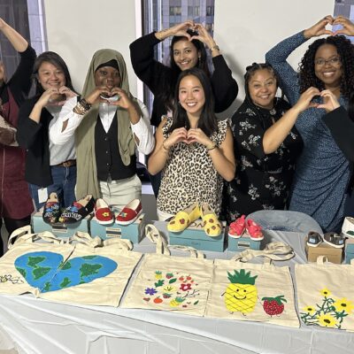 A group of smiling participants stands and makes heart gestures with their hands. They are displaying hand-painted TOMS shoes and tote bags created during the Paint Soles for Souls charitable team building event. thumbnail
