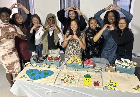A group of smiling participants stands and makes heart gestures with their hands. They are displaying hand-painted TOMS shoes and tote bags created during the Paint Soles for Souls charitable team building event.
