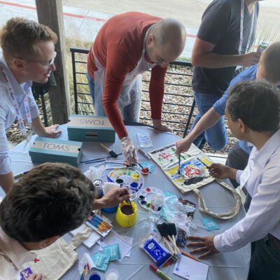 A group of participants collaborates on painting TOMS shoes and tote bags during the Paint Soles for Souls charitable team building event. The table is filled with art supplies, including paints, brushes, and TOMS shoeboxes, as participants creatively design items that will be donated. thumbnail