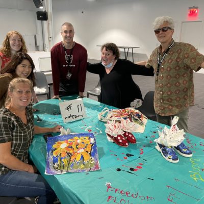 A group of participants poses around a table displaying their hand-painted TOMS shoes and decorated tote bags during the Paint Soles for Souls charitable team building event. The colorful items, featuring floral and abstract designs, will be donated to children in need in South America. The event encourages creativity, teamwork, and giving back to the community. thumbnail