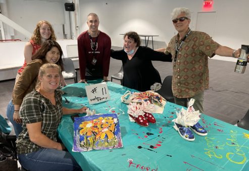A group of participants poses around a table displaying their hand-painted TOMS shoes and decorated tote bags during the Paint Soles for Souls charitable team building event. The colorful items, featuring floral and abstract designs, will be donated to children in need in South America. The event encourages creativity, teamwork, and giving back to the community.