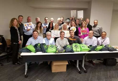 A group of people posing in a conference room behind a table filled with green drawstring bags. Some individuals are holding up supportive posters with words like 'Hope,' 'Stay Strong,' and 'You Are Cancer-Free.' The group is smiling, showing support for cancer patients as part of a team building event focused on creating care packages.