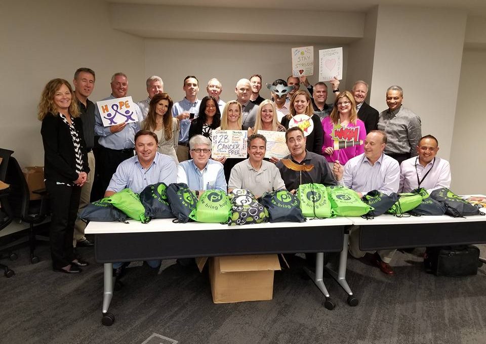 A group of people posing in a conference room behind a table filled with green drawstring bags. Some individuals are holding up supportive posters with words like 'Hope,' 'Stay Strong,' and 'You Are Cancer-Free.' The group is smiling, showing support for cancer patients as part of a team building event focused on creating care packages.