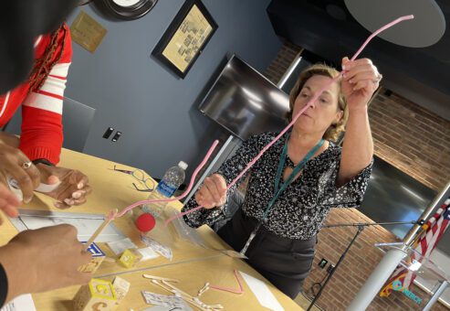 A woman carefully balancing a long pink pipe cleaner while participating in a hands-on team building challenge. She is surrounded by small wooden blocks, sticks, and other materials on a table.