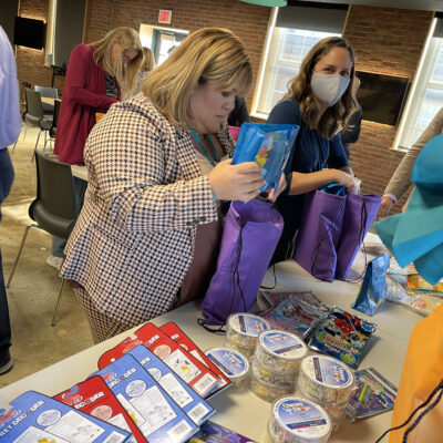 A group of people are participating in a team building activity where they are assembling care packages. A woman in the foreground is holding a purple bag and carefully placing an item inside. Various supplies are laid out on the table to put together care-kits to give to cancer patients thumbnail