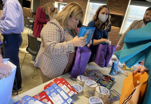 A group of people are participating in a team building activity where they are assembling care packages. A woman in the foreground is holding a purple bag and carefully placing an item inside. Various supplies are laid out on the table to put together care-kits to give to cancer patients