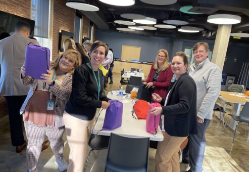 Group of smiling participants assembling colorful care packages for cancer patients during a TeamBonding event. The event, called Operation Cancer Care, combines fun team building activities with the meaningful goal of supporting cancer patients through the creation of care packages.