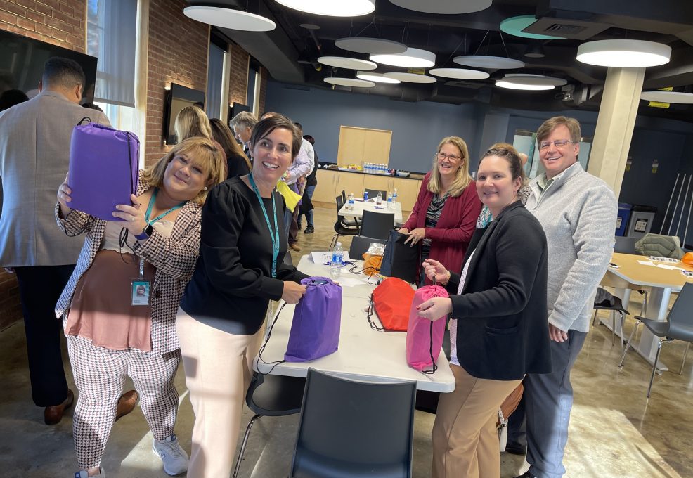 Group of smiling participants assembling colorful care packages for cancer patients during a TeamBonding event. The event, called Operation Cancer Care, combines fun team building activities with the meaningful goal of supporting cancer patients through the creation of care packages.