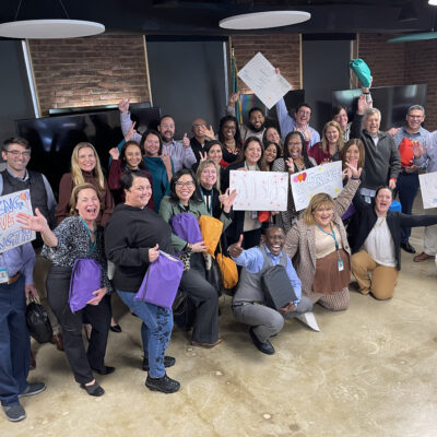 A large group of people posing together during a team building event. Some are holding colorful backpacks, while others hold hand-drawn supportive signs with messages like 'Sending Love & Strength.' The group is smiling, cheering, and making celebratory gestures, creating a positive and uplifting atmosphere. thumbnail