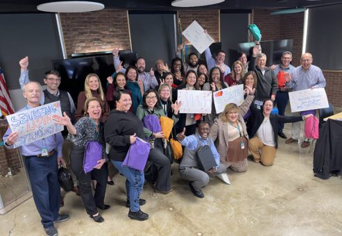A large group of people posing together during a team building event. Some are holding colorful backpacks, while others hold hand-drawn supportive signs with messages like 'Sending Love & Strength.' The group is smiling, cheering, and making celebratory gestures, creating a positive and uplifting atmosphere.