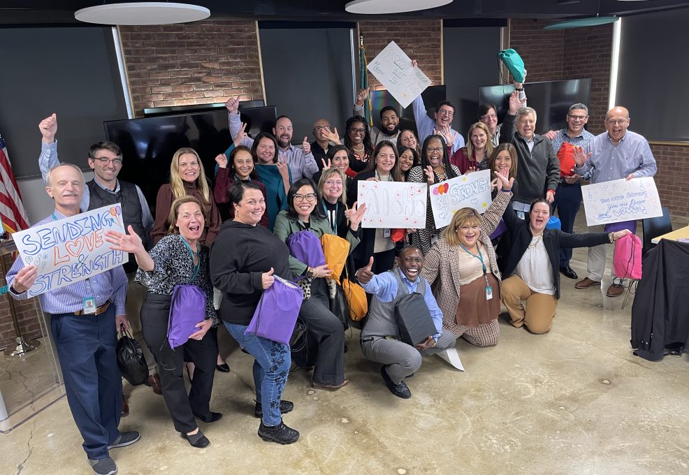 A large group of people posing together during a team building event. Some are holding colorful backpacks, while others hold hand-drawn supportive signs with messages like 'Sending Love & Strength.' The group is smiling, cheering, and making celebratory gestures, creating a positive and uplifting atmosphere.