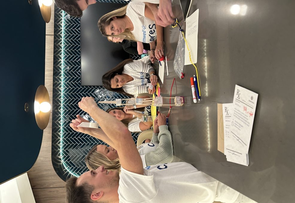 A group of people engage in a team building activity around a table. They are working together to build a structure using various materials such as string and sticks, with a focused and collaborative atmosphere.