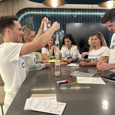 A group of people engage in a team building activity around a table. They are working together to build a structure using various materials such as string and sticks, with a focused and collaborative atmosphere. thumbnail
