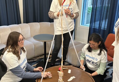 Three team members are participating in a team building challenge. They are gathered around a small round table, constructing a tall tower using wooden sticks, tape, and string. Two participants are sitting on the floor working on the base, while one stands, adjusting the top of the structure.