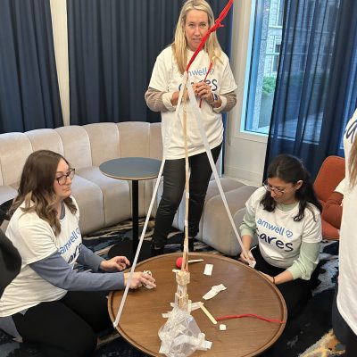 Three team members are participating in a team building challenge. They are gathered around a small round table, constructing a tall tower using wooden sticks, tape, and string. Two participants are sitting on the floor working on the base, while one stands, adjusting the top of the structure. thumbnail