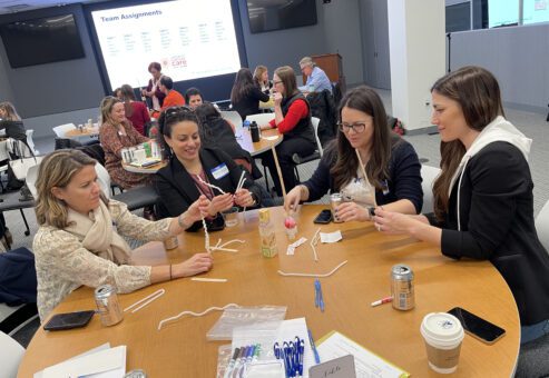 Four women are seated at a round table, engaging in a hands-on team building activity. They are using pipe cleaners, blocks, and small materials to build a structure. The room is filled with other groups participating in similar activities. A large screen in the background displays 'Team Assignments' and the event name, 'Operation Cancer Care.' The atmosphere is collaborative and focused, with various materials like markers, pens, and drinks on the table.