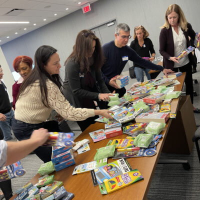 A group of individuals gather around a table filled with various school supplies, including pencils, erasers, and notebooks. They are sorting and organizing the supplies, as part of a team building CSR activity aimed at creating care packages for cancer patients. thumbnail